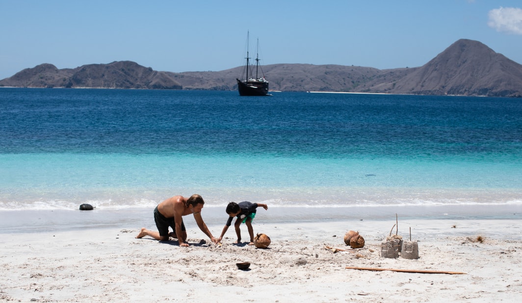A father and son building a sandcastle on a white beach, Dunia Baru at anchor in the bay behind