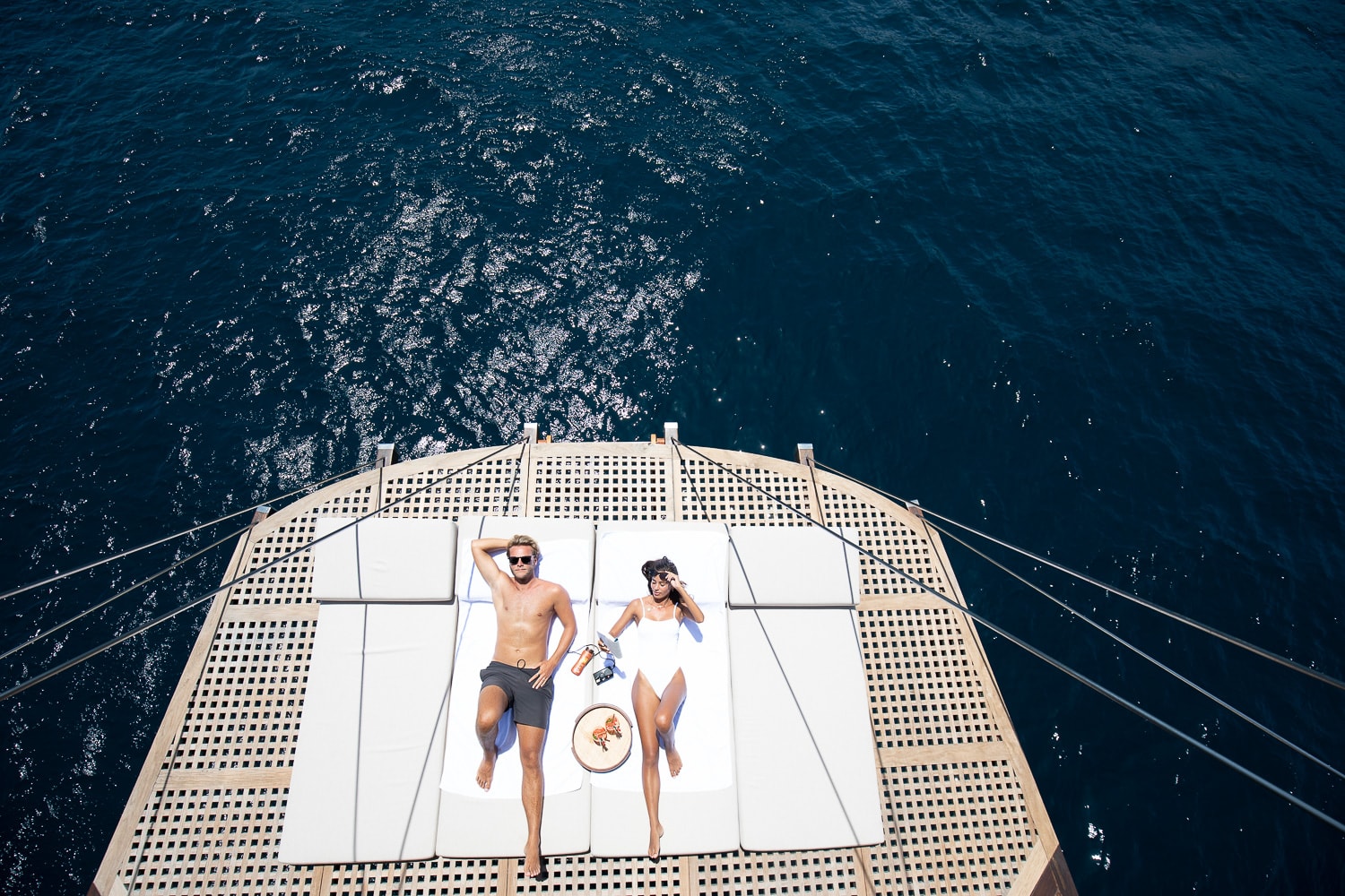 Two guests sunbathing on the netting at Dunia Baru's bow, open water below them, seen from the mast