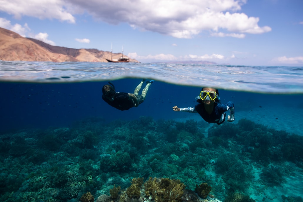 Two guests snorkelling over a shallow coral reef, Dunia Baru anchored in the bay beyond