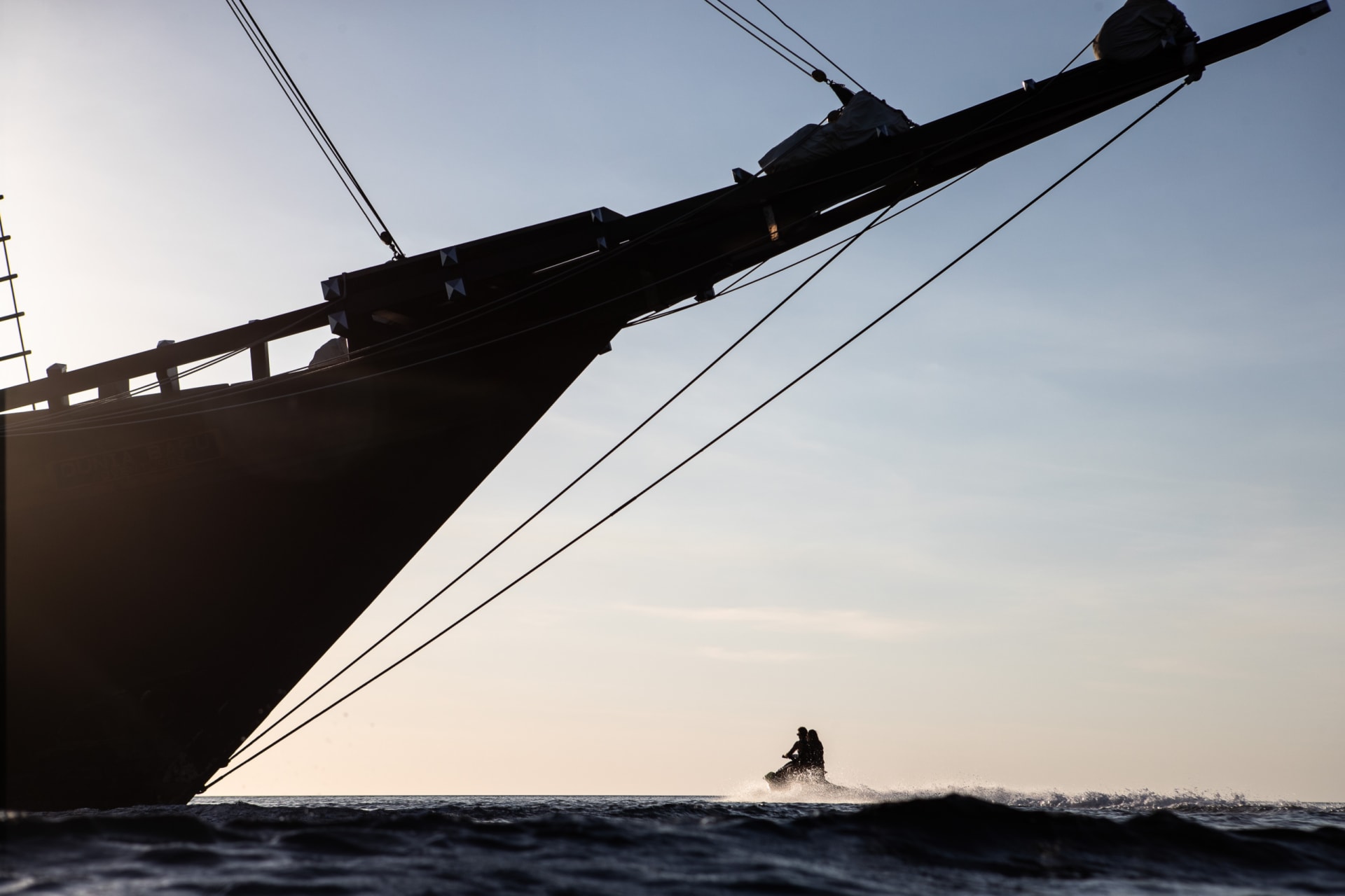 A jet ski crossing the water at dusk beneath Dunia Baru's bowsprit and rigging